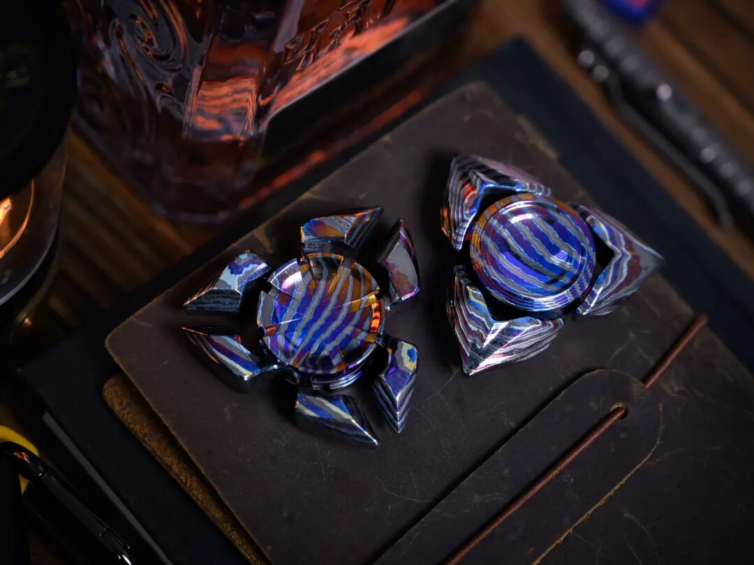 Two three-arm Zircuti spinners on a brown surface, with gold and blue bands inside different outlines