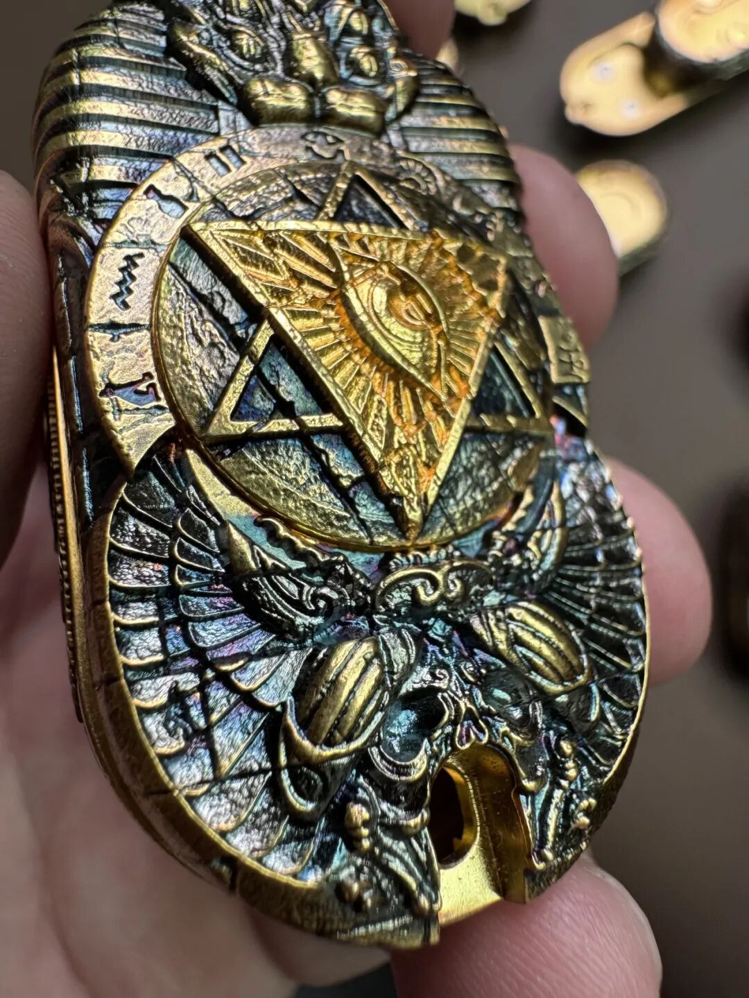 A Horus Eye slider held in a hand, with gold-coloured raised details against dark recessed decoration.