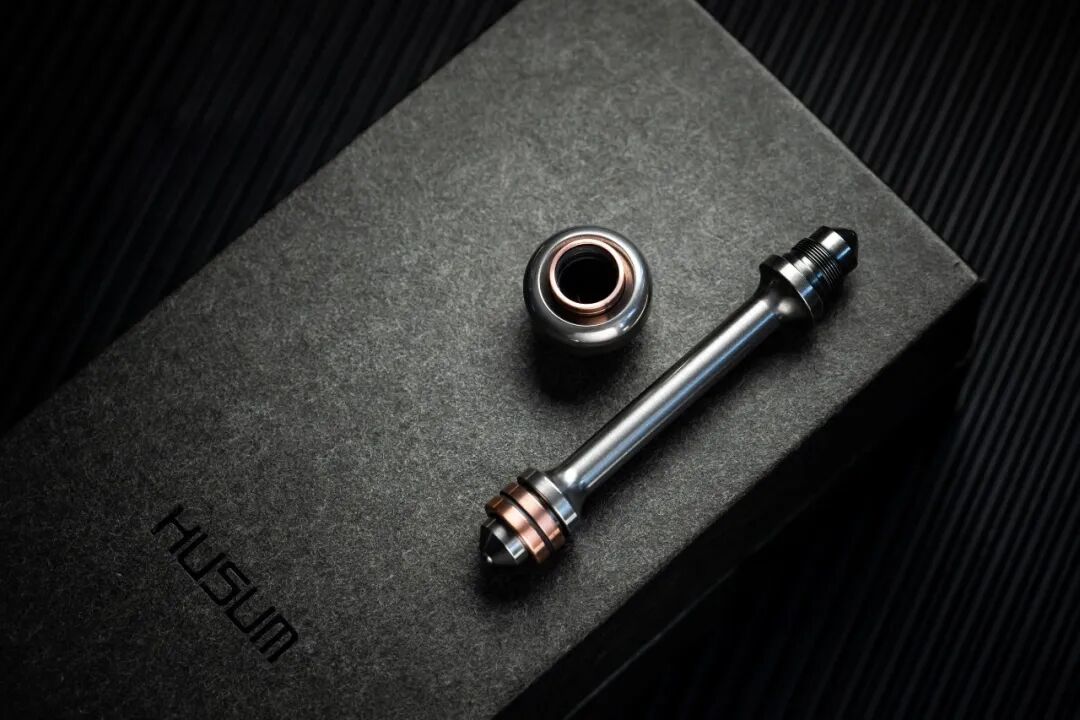 A stainless-steel connecting rod and adapter on a grey surface, with copper-coloured rings at the opening and end